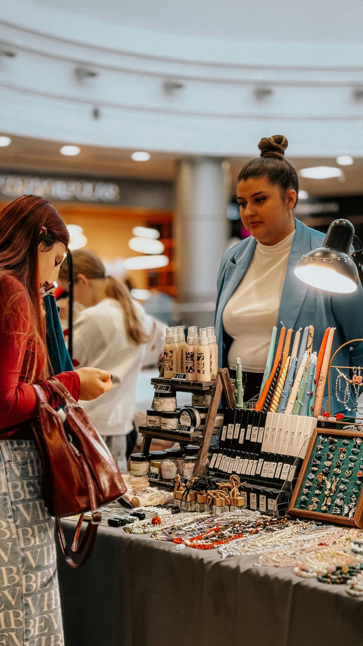 Women browsing handmade jewelry at indoor market stall.